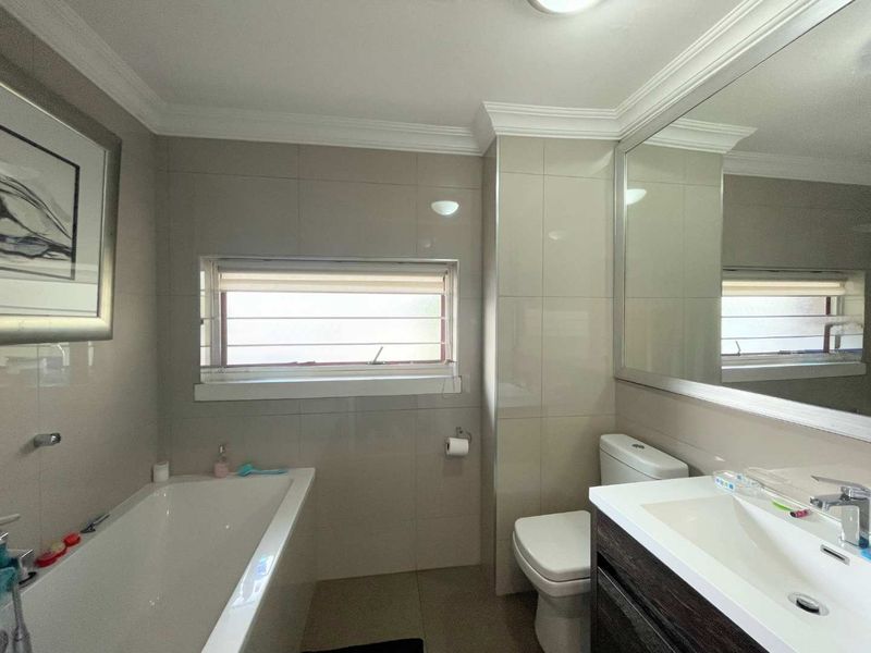 A well-maintained bathroom featuring a built-in bathtub, modern vanity with basin, large wall mirror, and neutral tiled finishes.