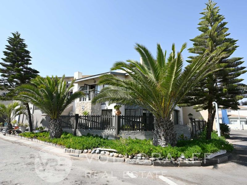 Front elevation of a residential property featuring stone cladding, a balcony, and a well-maintained garden with mature palm trees.