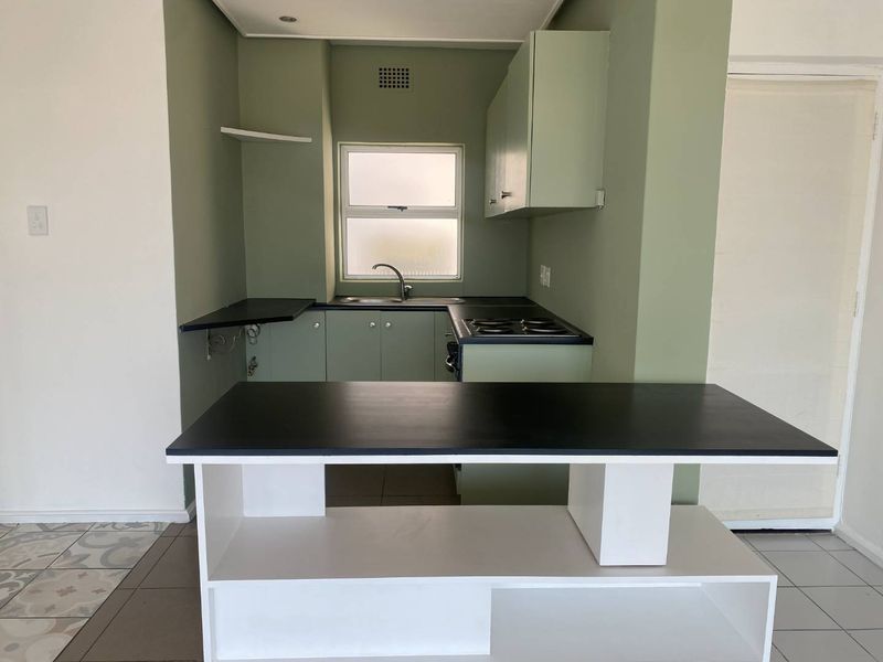 A compact kitchen featuring sage green cabinetry, black countertops, a built-in electric hob, and a stainless steel sink, offering a functional layout.