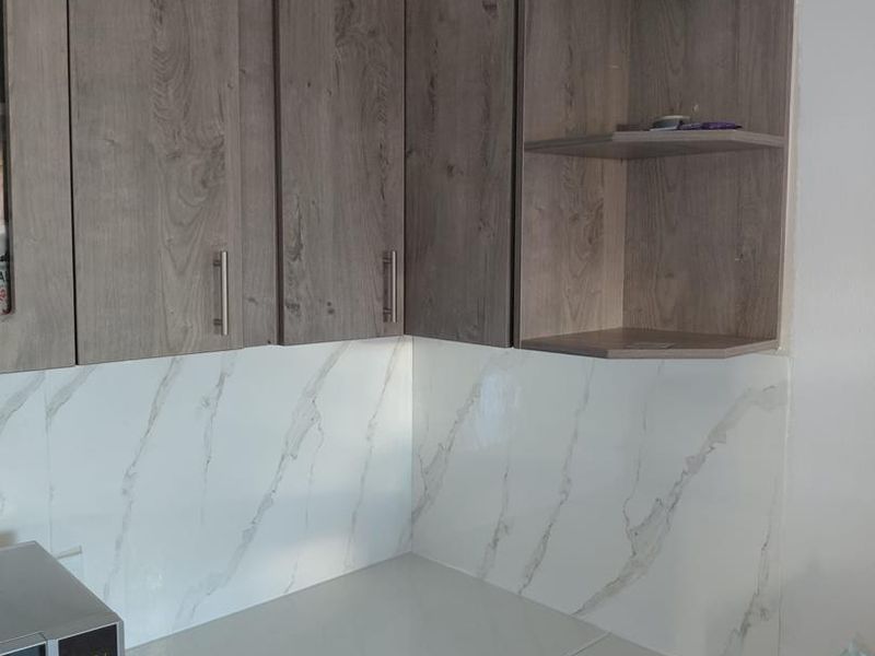 A modern kitchen corner featuring wood-grain laminate cabinetry, a marble-effect splashback, and clean stone-look countertops with an integrated microwave.