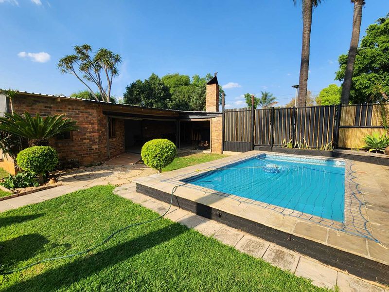 Outdoor pool area featuring a swimming pool with safety net, paved surround, and a covered brick built-in braai area in the garden.