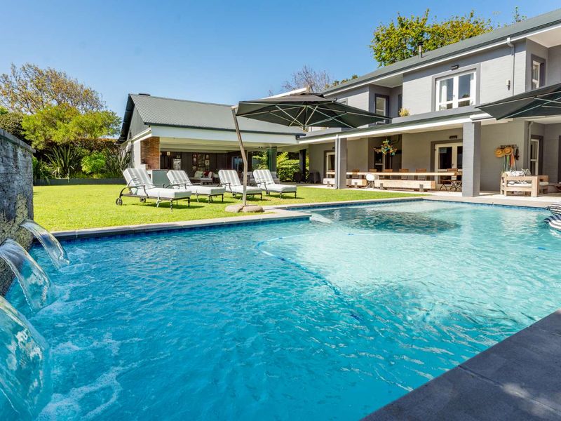 A well-maintained outdoor pool area featuring a modern swimming pool with water features, a spacious lawn, and a covered patio area.