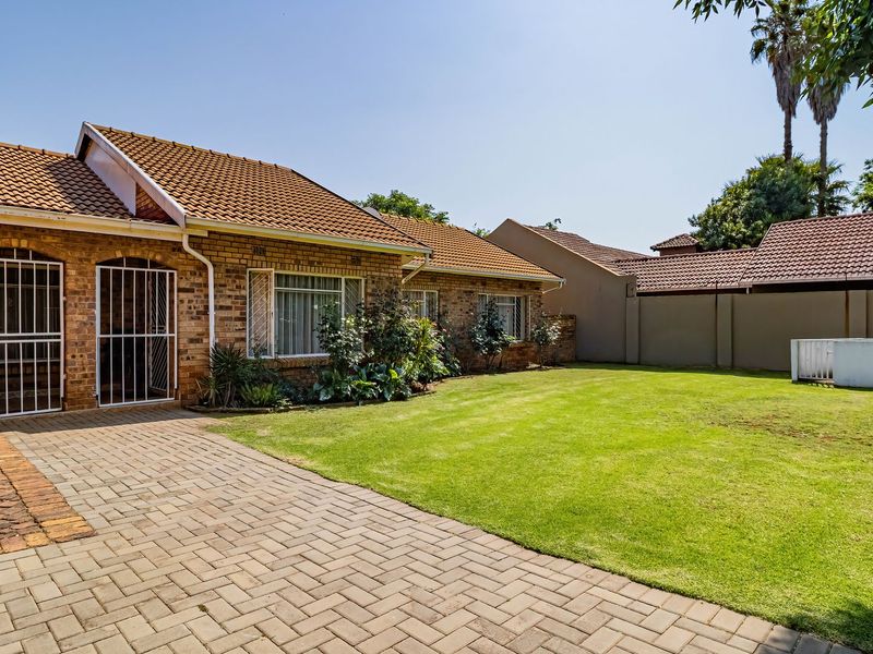 A well-maintained rear exterior view featuring a brick facade, tiled roof, paved patio area, and a spacious lawn enclosed by a boundary wall.