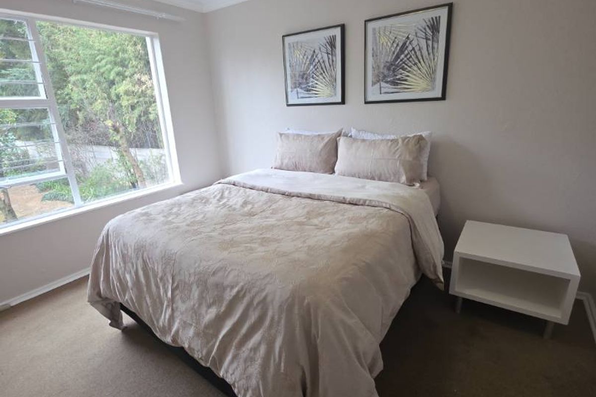 A well-lit bedroom featuring neutral wall tones, carpeted flooring, and a large window providing natural light.