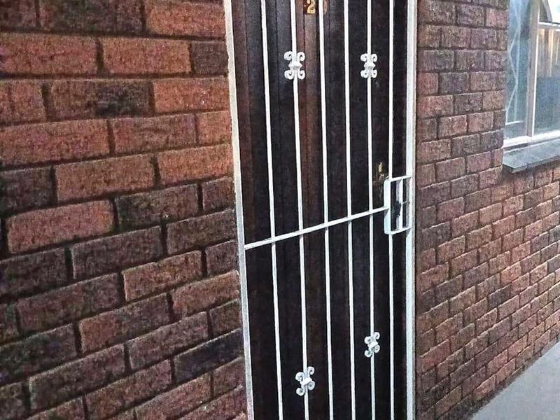 Exterior view of an apartment entrance featuring a solid door protected by a white metal security gate set within a face-brick wall.