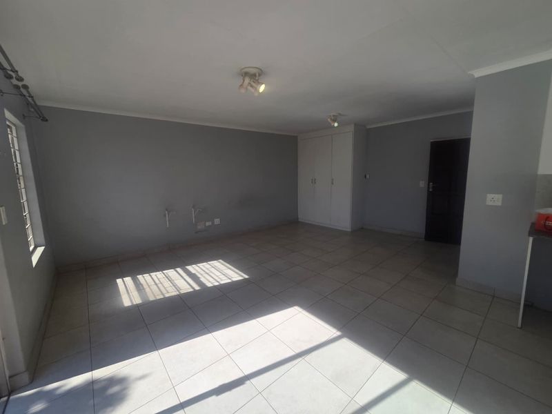 An open-plan living area featuring neutral grey walls, tiled flooring, built-in storage cupboards, and natural light through window security bars.