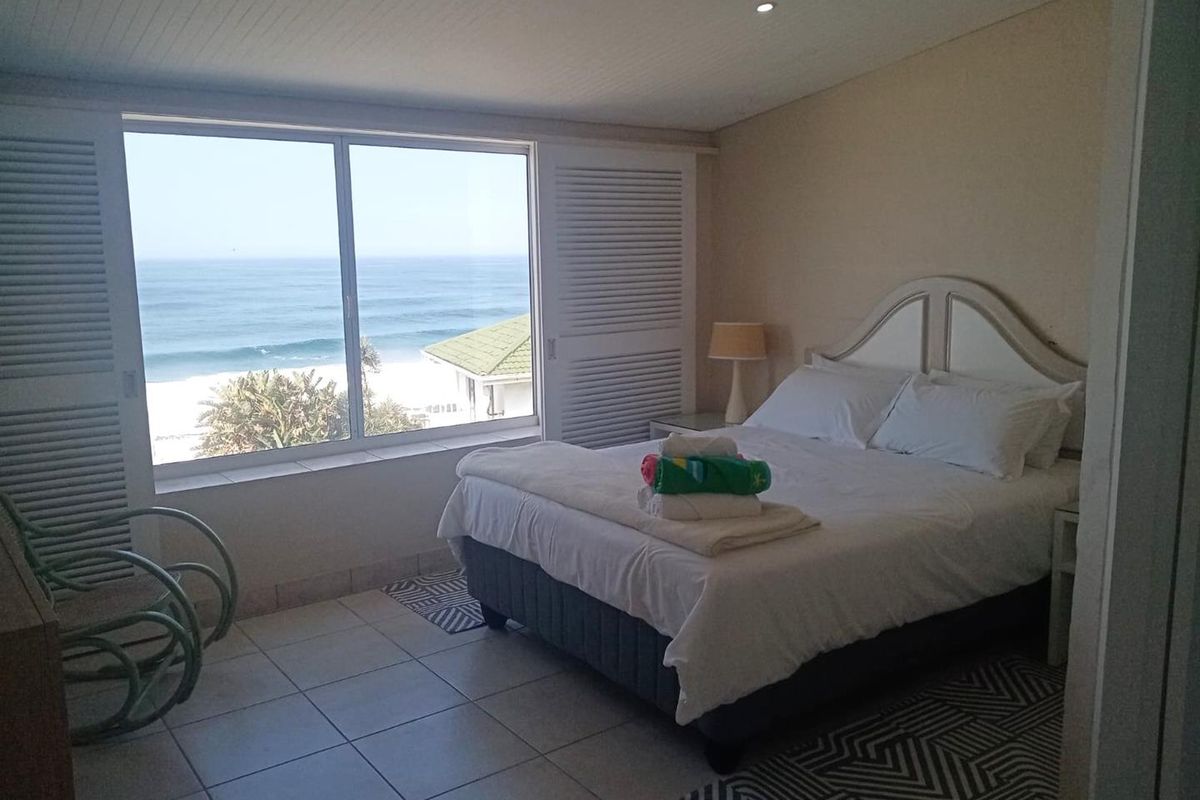 A bright bedroom featuring tiled flooring, recessed lighting, and louvered shutters, offering a direct view of the ocean.