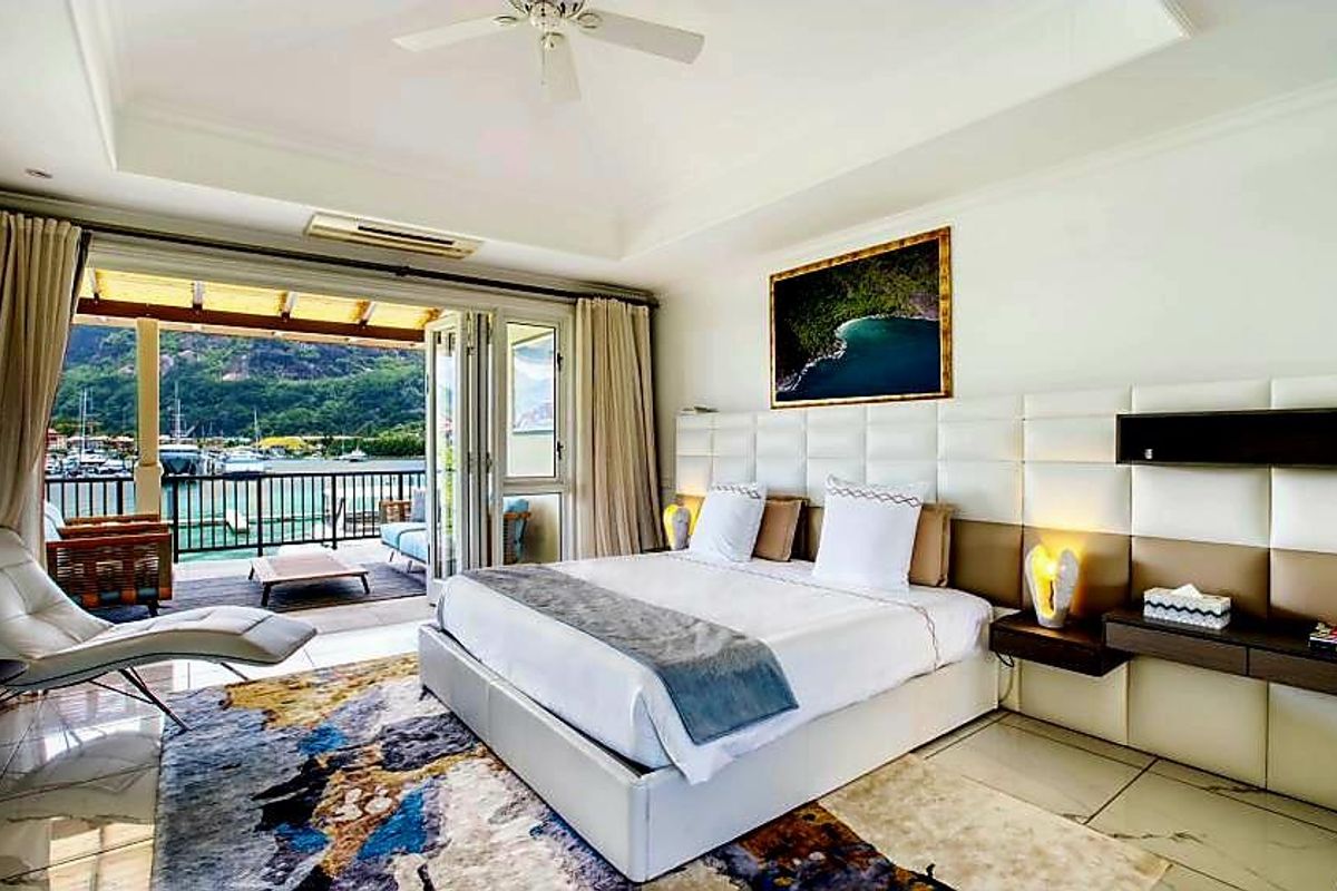 Spacious bedroom featuring a large upholstered headboard, floating bedside tables, ceiling fan, and bi-fold doors opening onto a balcony with marina views.
