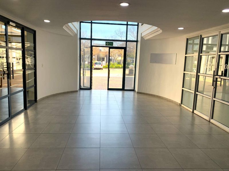 A bright and spacious office building entrance lobby featuring tiled flooring, recessed ceiling lighting, and large glass panel doors leading to the exterior.