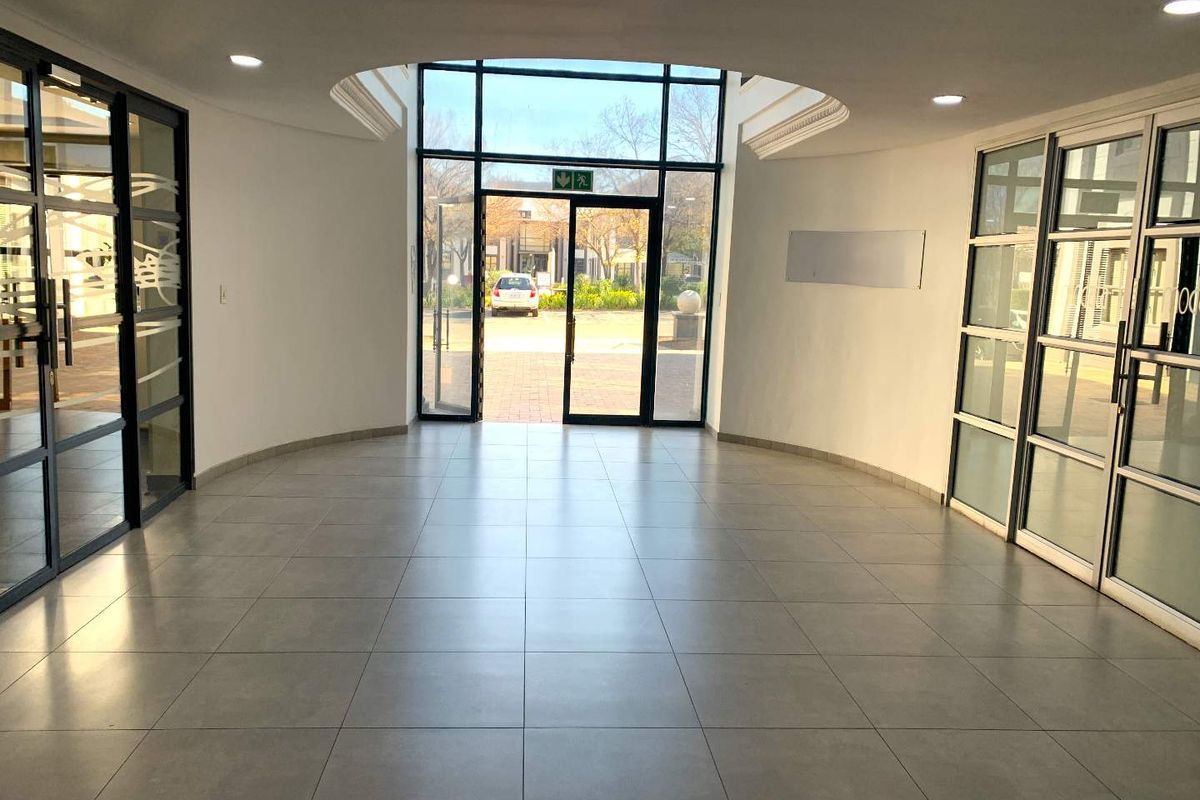A bright and spacious office building entrance lobby featuring tiled flooring, recessed ceiling lighting, and large glass panel doors leading to the exterior.