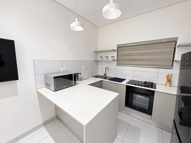 Modern kitchen featuring sleek quartz countertops, built-in oven, gas hob, stainless steel sink, and minimalist floating shelving with neutral tiled finishes.