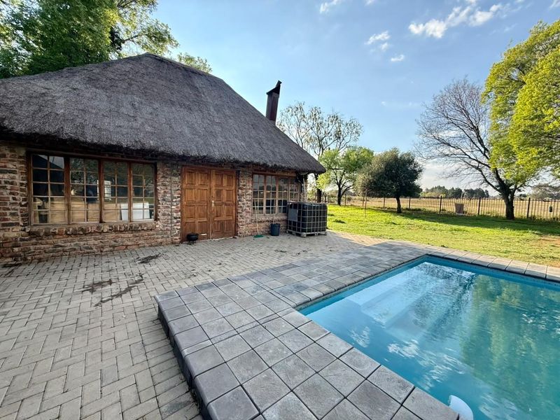 Exterior view of a rustic farmhouse with a thatched roof, featuring a paved patio area and a swimming pool set in a spacious garden.