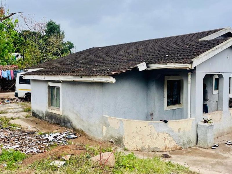 Exterior view of a single-story house showing significant wear to the facade, roof tiles, and guttering, with debris present in the surrounding yard.