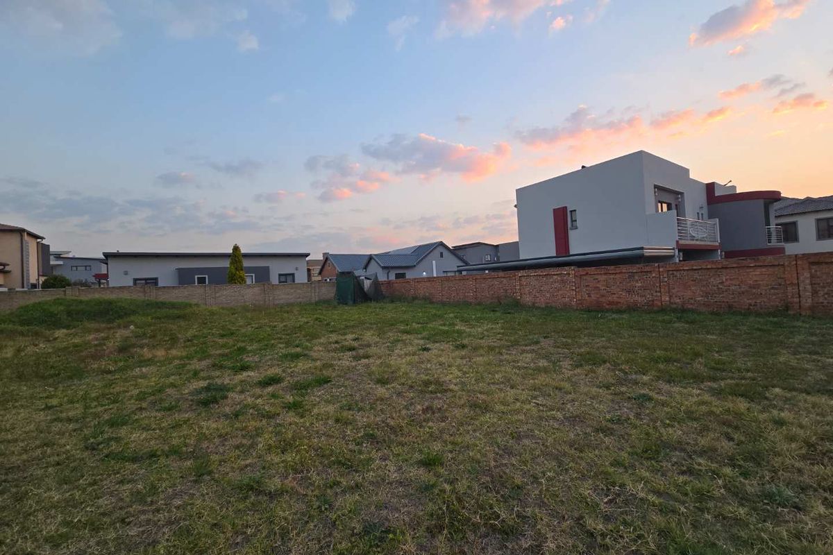 A vacant residential plot featuring a grass lawn and a brick perimeter wall, situated in a developed neighborhood.
