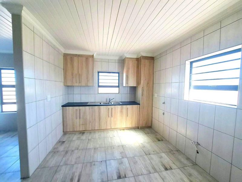 A clean, empty kitchen featuring light wood-look cabinetry, tiled walls and flooring, and a functional stainless steel sink with louver windows.