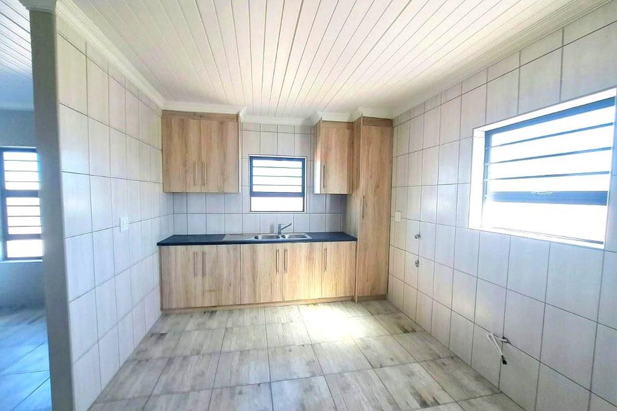 A clean, empty kitchen featuring light wood-look cabinetry, tiled walls and flooring, and a functional stainless steel sink with louver windows.