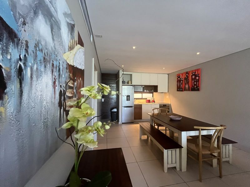 An open-plan living and dining area featuring tiled flooring, recessed lighting, and a compact, functional kitchen with built-in cabinetry and appliances.