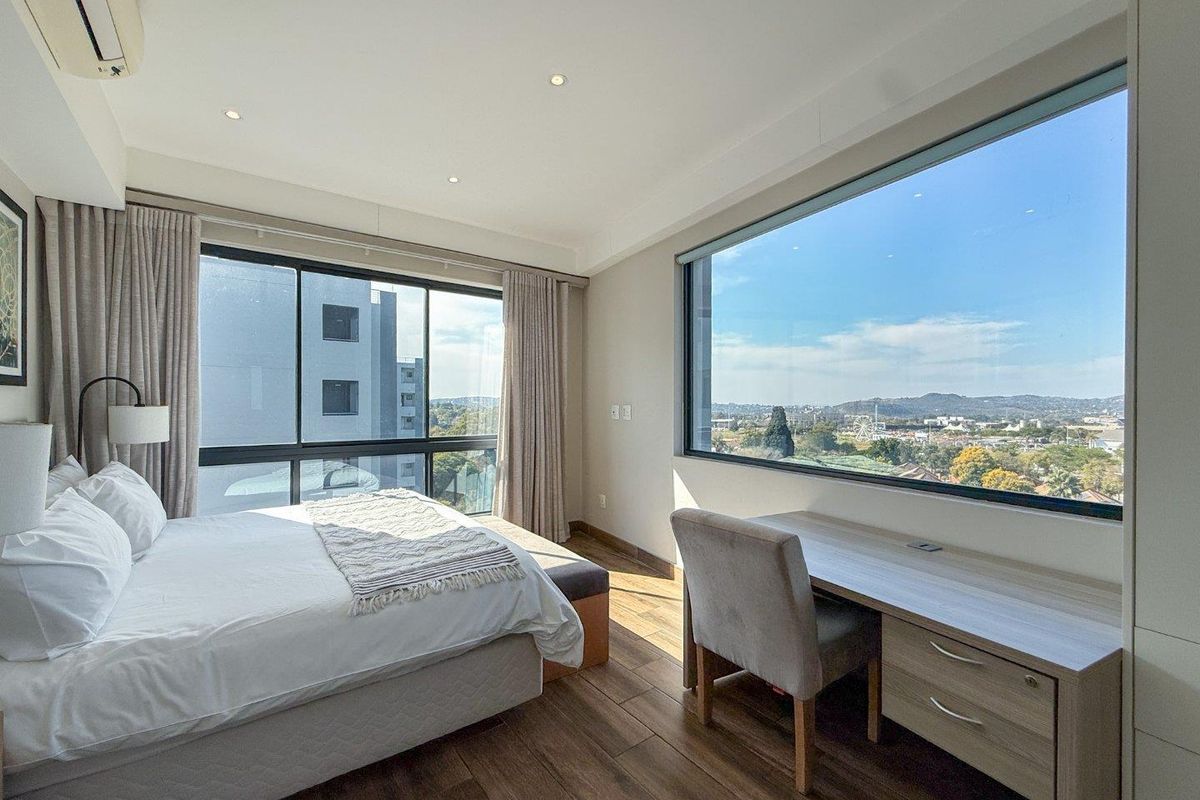 A bright and modern bedroom featuring large windows with panoramic views, wood-look flooring, and a built-in desk area.