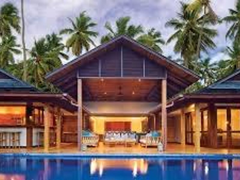 A stunning villa exterior featuring a central covered patio, a sparkling swimming pool, and a tropical garden setting with palm trees.