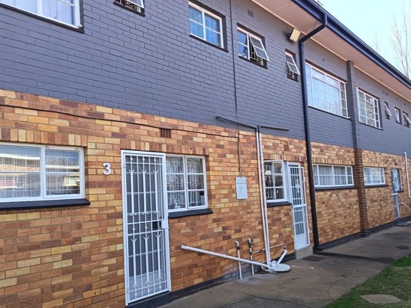 Exterior view of an apartment building featuring a two-tone brick facade, security gates, and multiple casement windows along a concrete walkway.