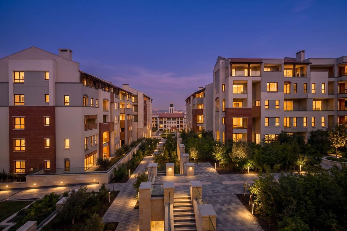 A beautifully illuminated evening view of the apartment complex, featuring landscaped courtyards, paved walkways, and modern architectural facades.