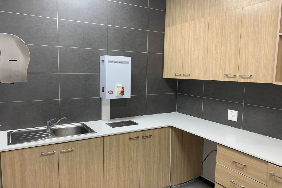 A modern office kitchenette featuring light wood cabinetry, white countertops, a stainless steel sink, and a wall-mounted water boiler.