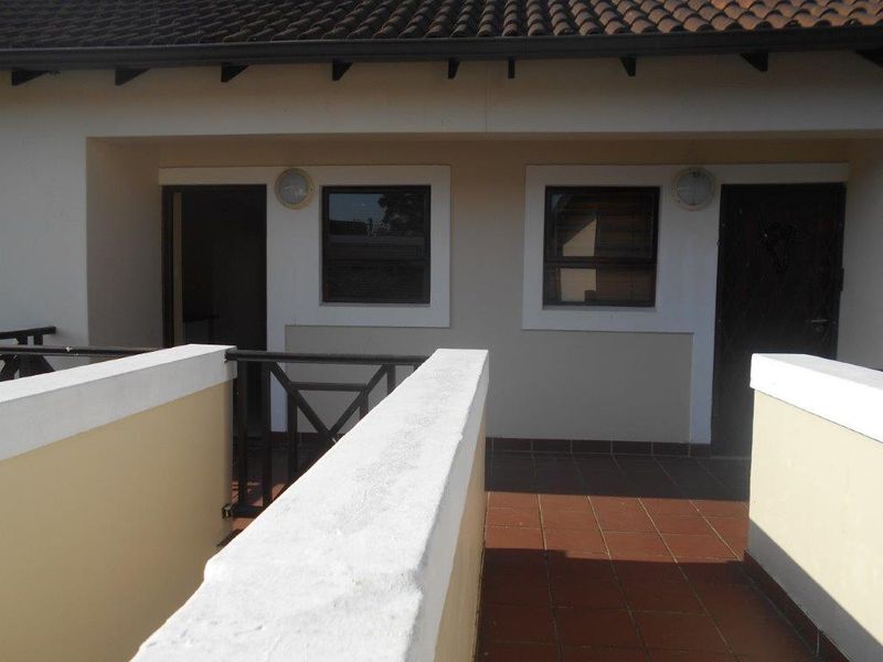 A covered balcony area featuring tiled flooring, a protective balustrade, and exterior wall lighting, providing access to the apartment entrance.