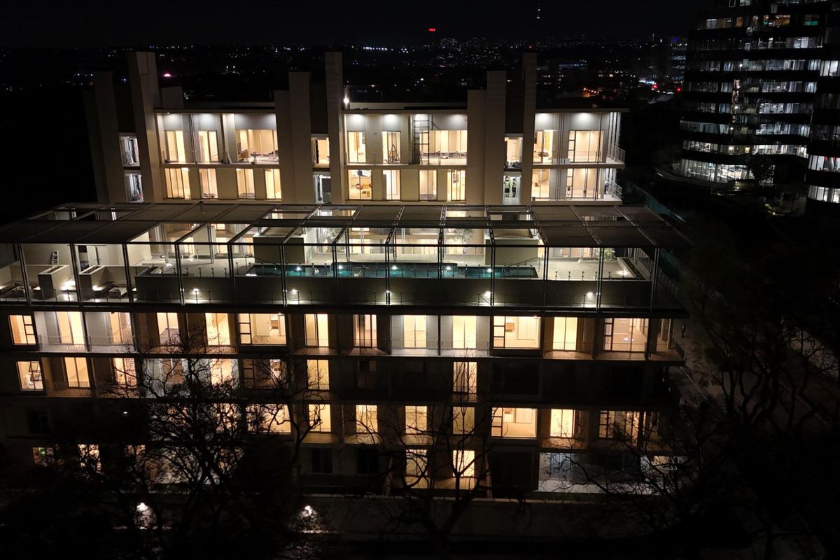 Nighttime aerial view of a modern multi-story apartment complex featuring a rooftop swimming pool and illuminated balconies.