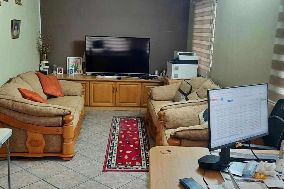 A living room featuring tiled flooring, built-in cabinetry, and roller blinds, currently utilized as a multi-purpose living and workspace area.