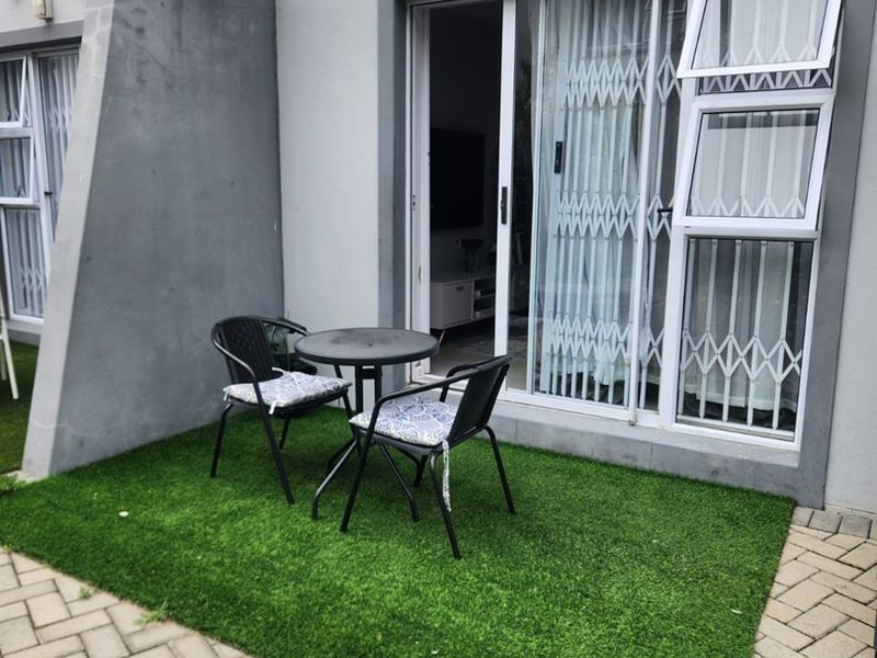 A private patio area featuring artificial grass, paved surround, and a sliding glass door with security gate, sheltered by a structural pergola.