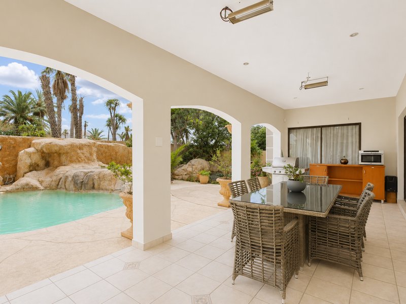 Covered patio area featuring tiled flooring, arched architectural details, and integrated outdoor heaters, overlooking a landscaped swimming pool with a natural rock feature.