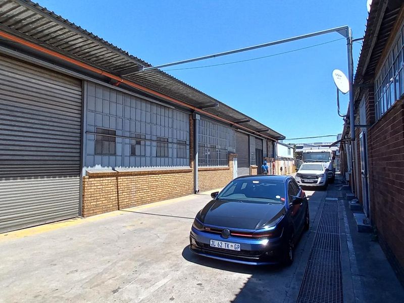 Exterior view of an industrial warehouse facility featuring brick walls, large roller shutter doors, and steel-framed windows along a concrete driveway.