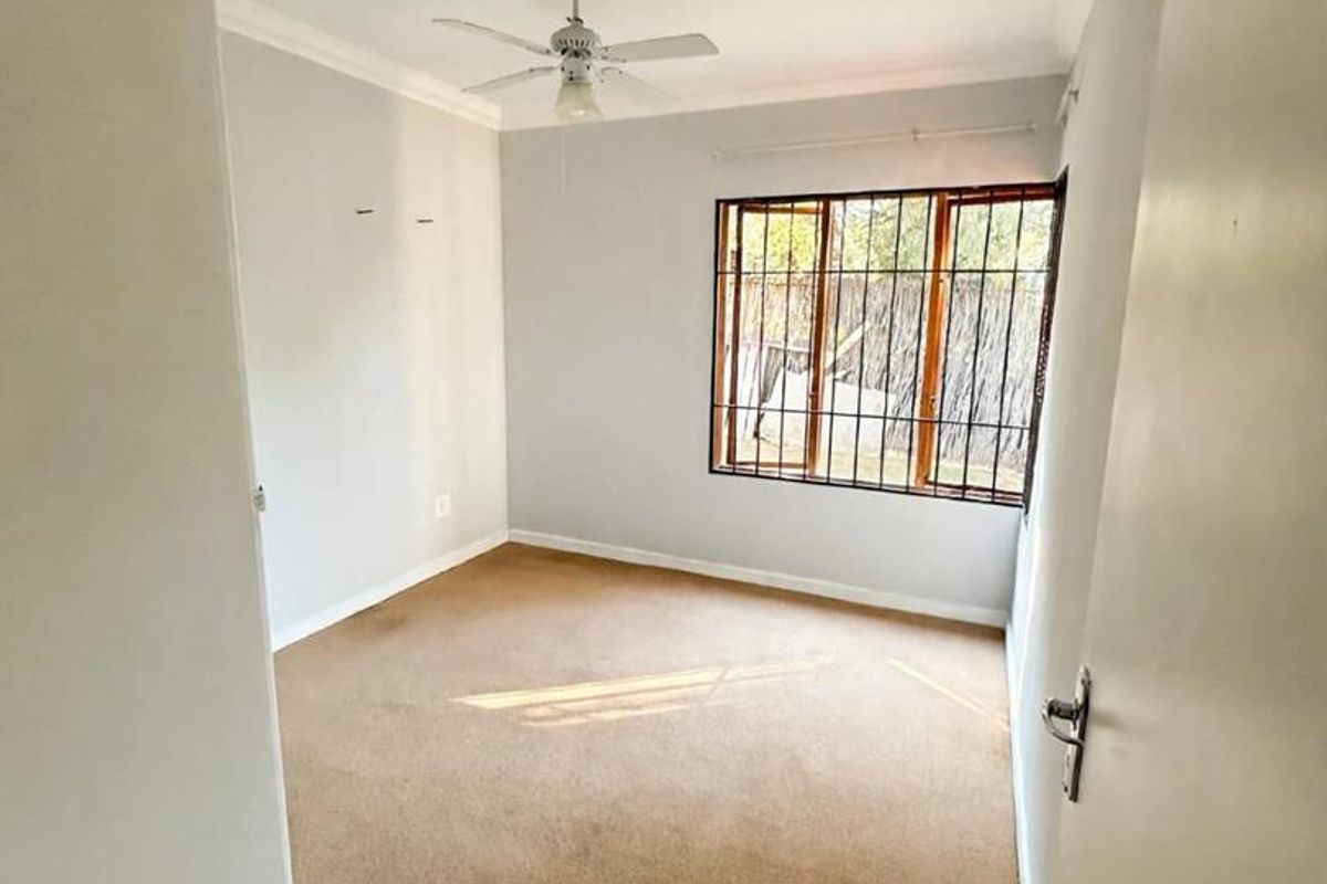 A vacant bedroom featuring neutral wall paint, carpeted flooring, a ceiling fan, and a window fitted with security bars.