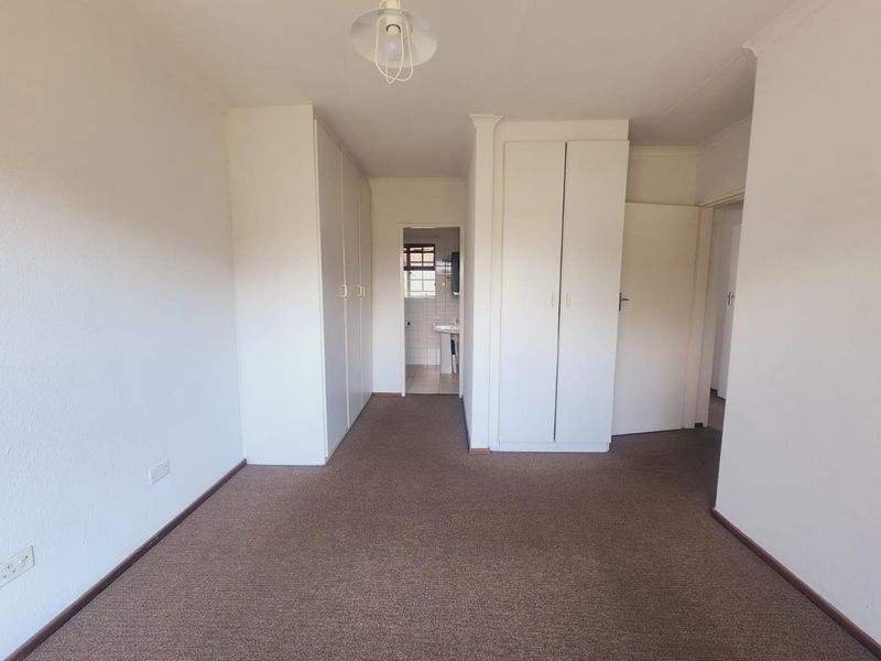 A spacious, empty bedroom featuring neutral walls, carpeted flooring, and ample built-in cupboard storage, with an adjoining view into a bathroom area.
