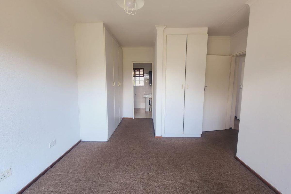 A spacious, empty bedroom featuring neutral walls, carpeted flooring, and ample built-in cupboard storage, with an adjoining view into a bathroom area.