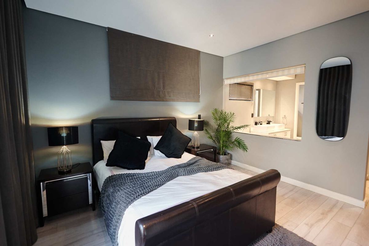 A modern bedroom featuring light wood-look flooring, recessed ceiling lighting, and an open-plan design with a view into the adjoining en-suite bathroom.