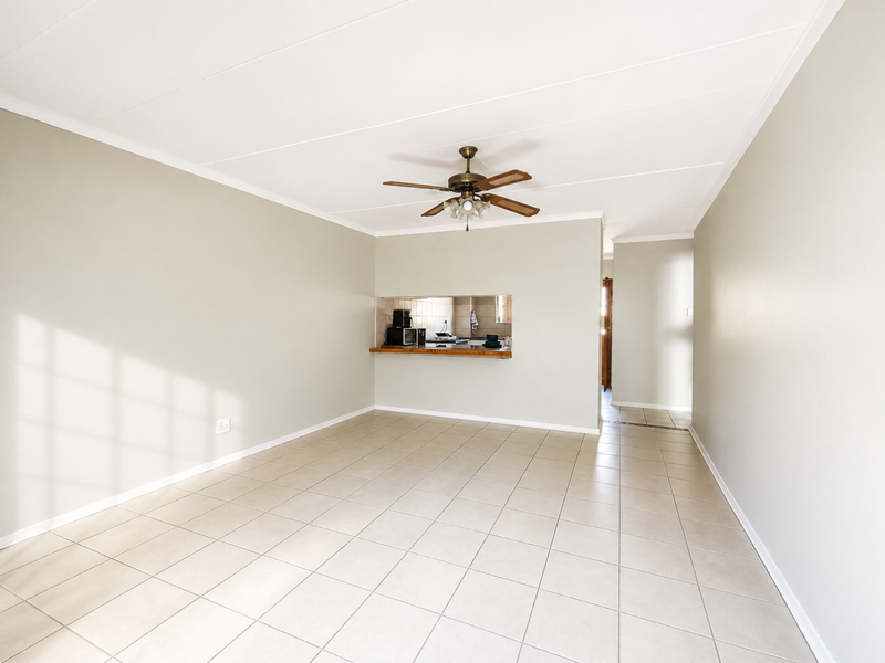 A spacious, empty living area featuring neutral tiled flooring, a ceiling fan, and a convenient serving hatch connecting to the kitchen.