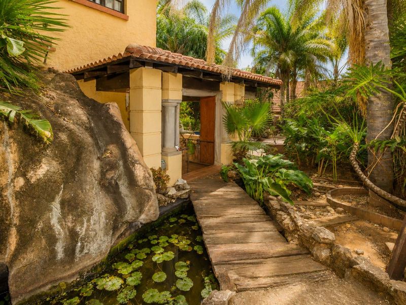 A rustic rear garden entrance featuring a wooden footbridge over a small pond, set against a stone rock face and lush tropical landscaping.