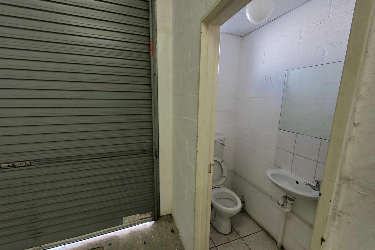 A functional industrial restroom featuring a toilet, wall-mounted basin, and mirror, located adjacent to a large roller shutter door.