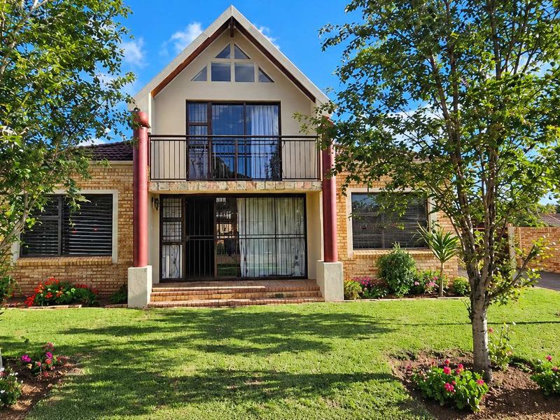 Front elevation of a double-story townhouse featuring a brick facade, a balcony with metal railings, and a well-maintained front garden with a paved driveway.