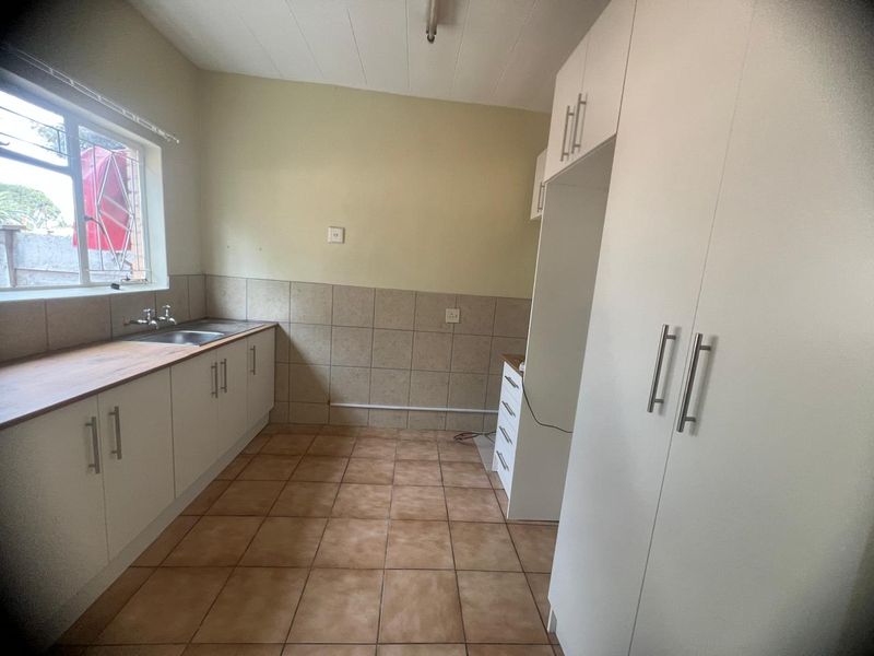 A clean, empty kitchen featuring white built-in cabinetry, tiled splashbacks, a stainless steel sink, and tiled flooring with ample storage space.