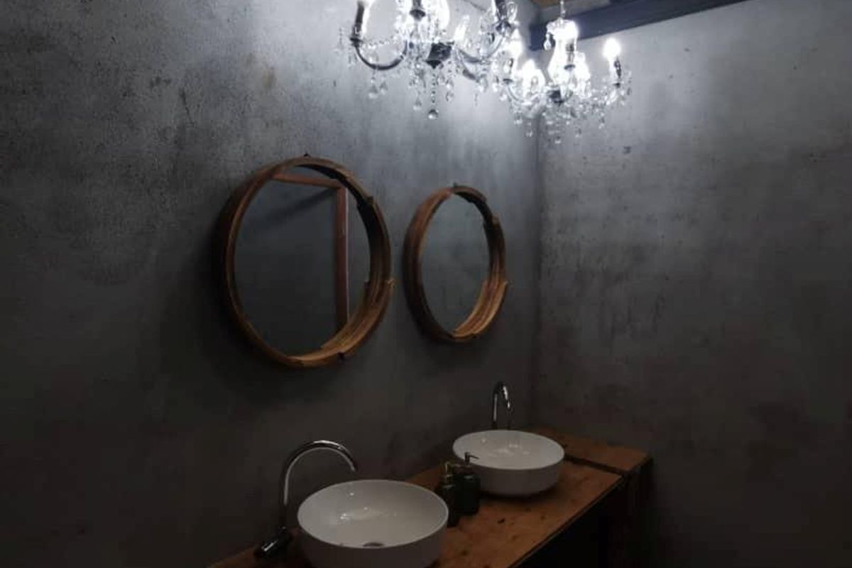 A modern industrial-style bathroom featuring a double vanity with vessel sinks, concrete-textured walls, and exposed timber ceiling beams with a crystal chandelier.