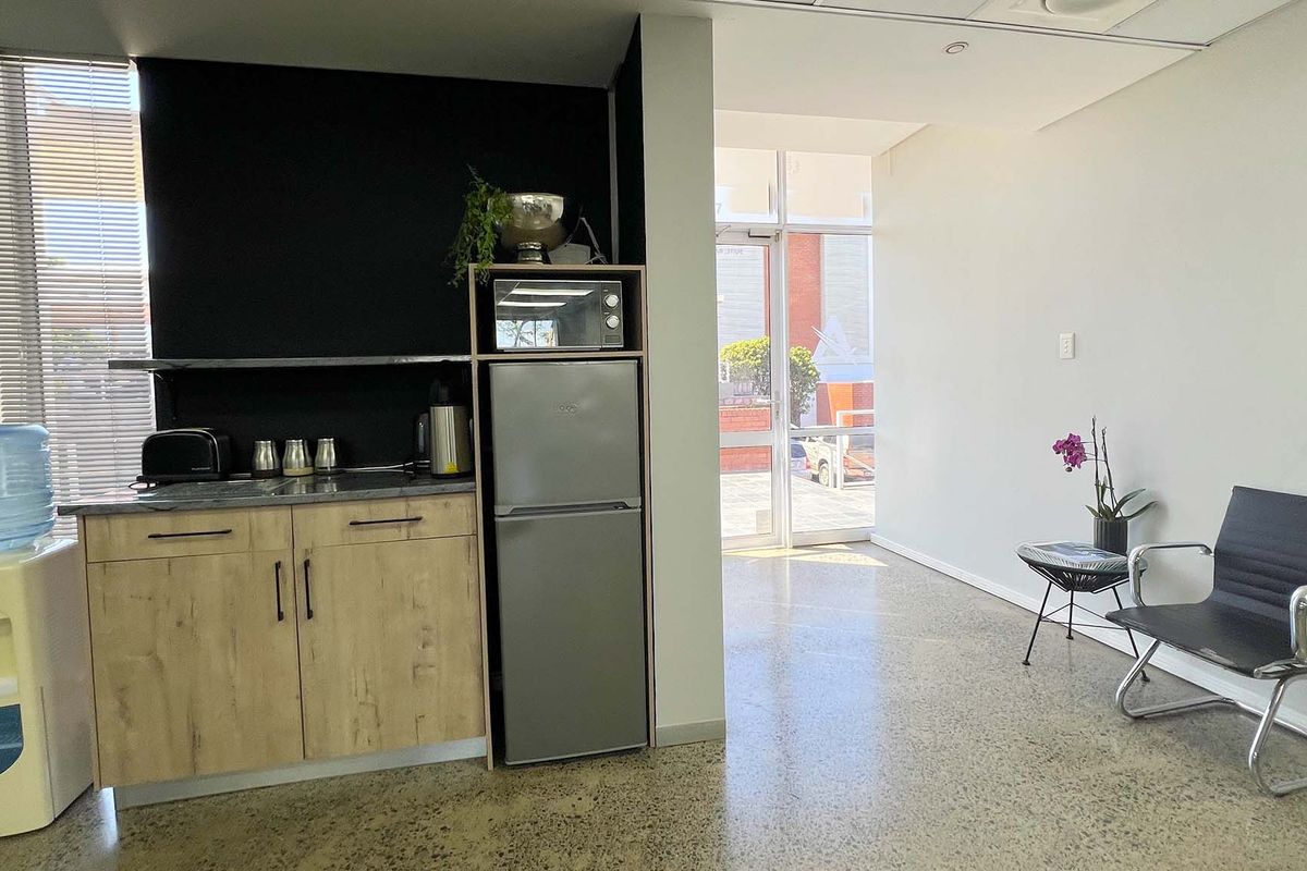 A compact office kitchenette featuring wood-look cabinetry, a stone countertop, and polished concrete flooring, leading to a bright entrance area.