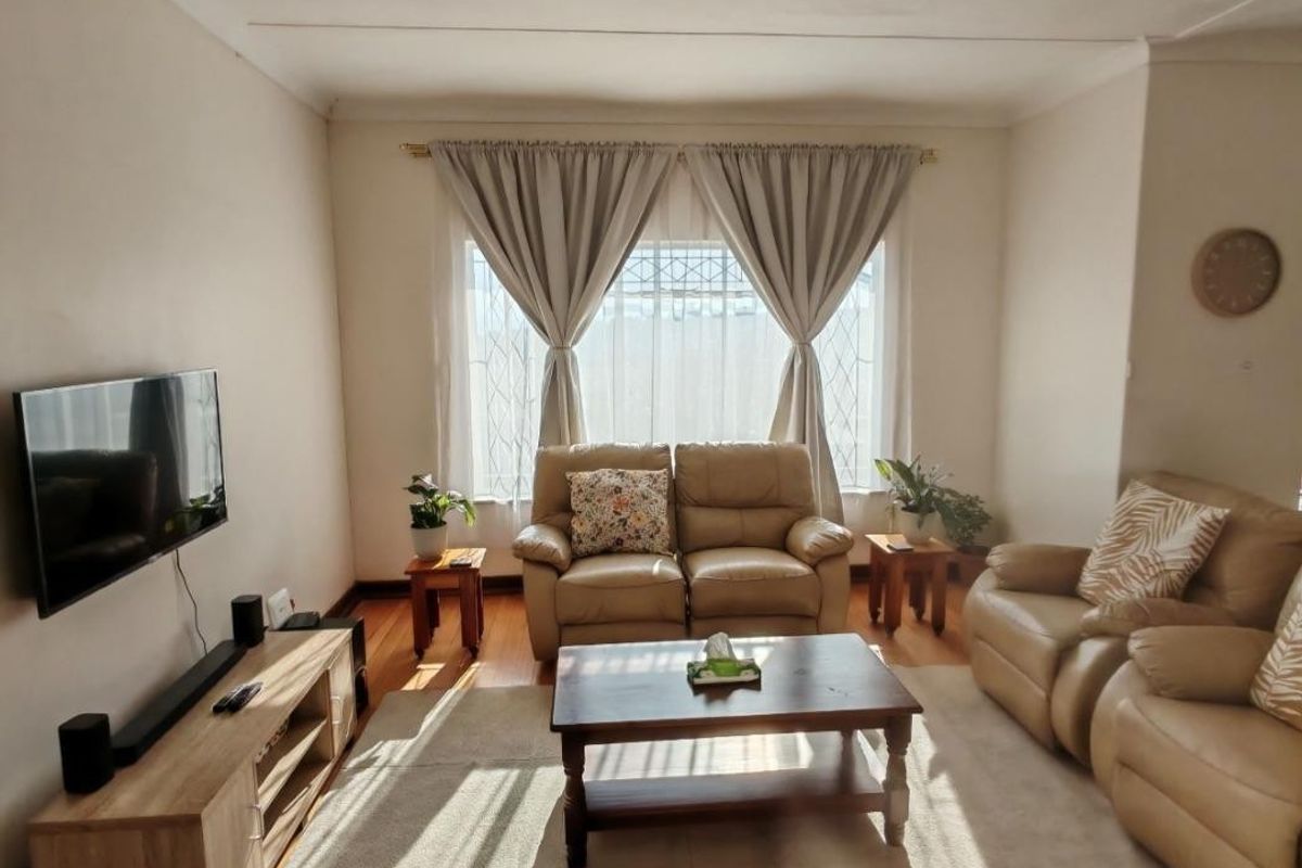 A comfortable living room featuring warm wooden flooring, natural light through a large window with security gate, and neutral wall finishes.