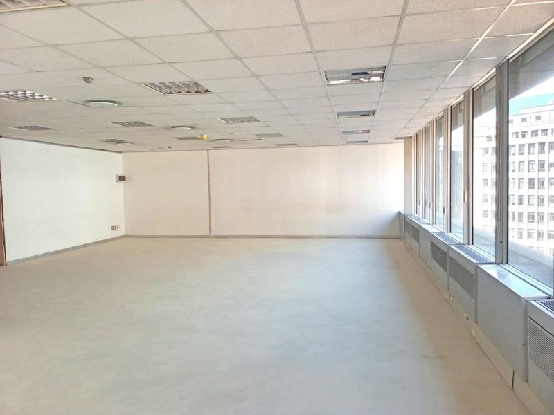 A spacious, empty office area featuring a suspended ceiling, recessed lighting, and large windows providing ample natural light and city views.