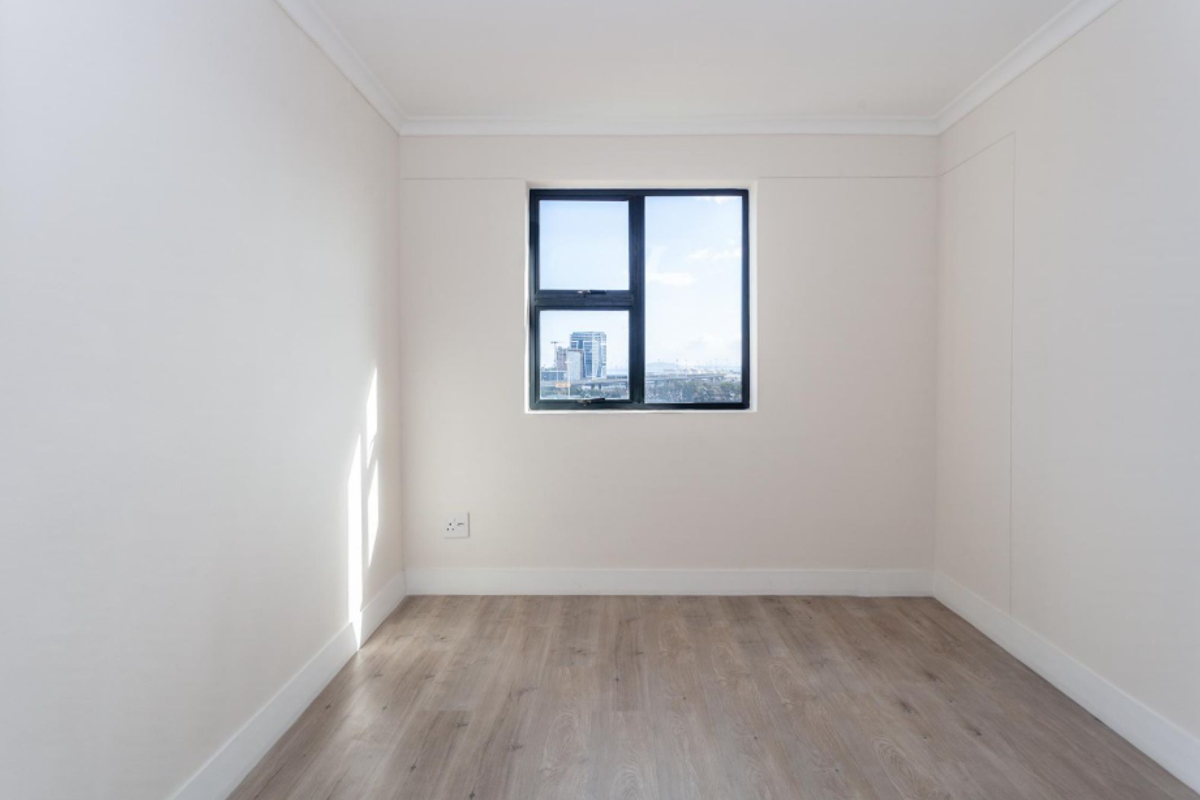A bright, empty bedroom featuring modern light wood-look flooring, neutral walls, and a window with a city view.