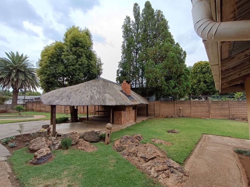 Spacious outdoor entertainment area featuring a traditional thatched lapa with a built-in brick braai, set within a landscaped garden with mature trees.