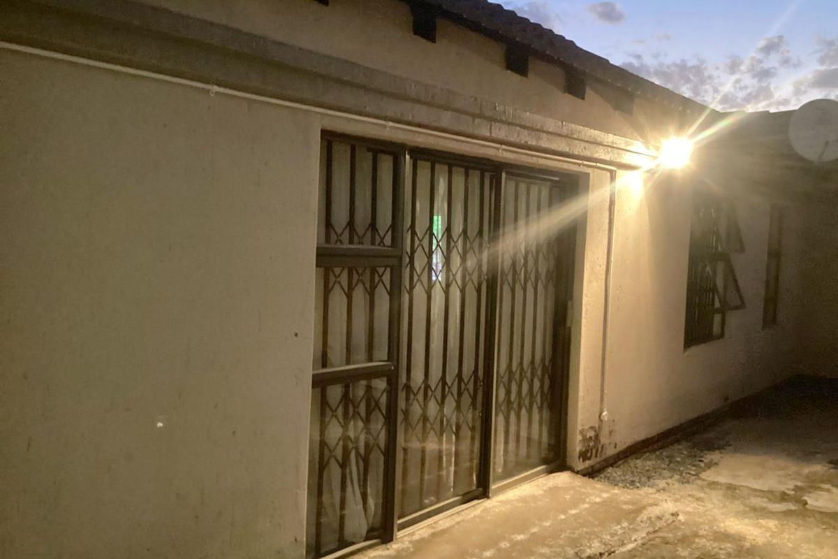 Rear exterior view of the property featuring sliding glass doors with security gates, concrete paving, and exterior wall lighting.