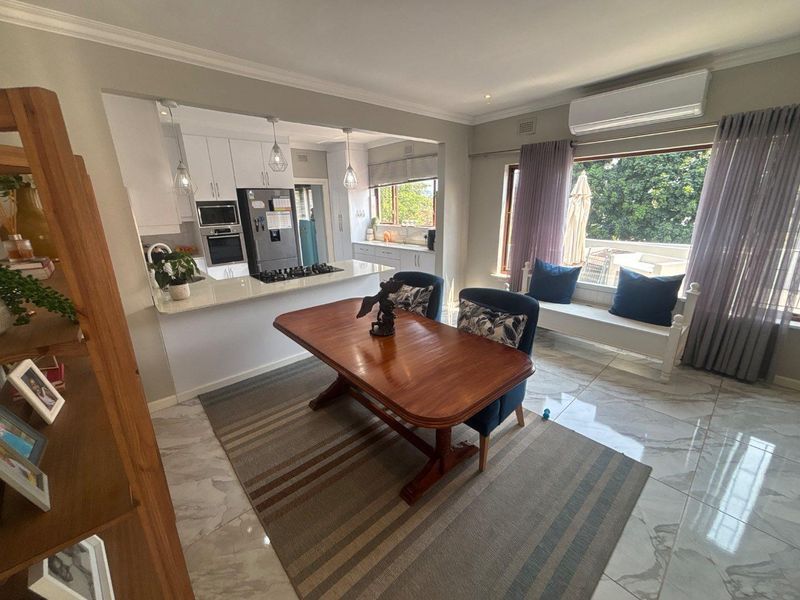 Open-plan kitchen and dining area featuring modern white cabinetry, stone countertops, integrated appliances, and polished tiled flooring with abundant natural light.
