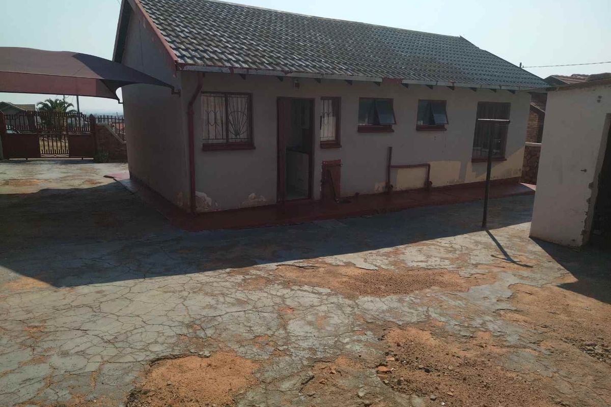 Rear view of the property featuring a tiled roof, security bars on windows, and a large concrete driveway area in need of maintenance.